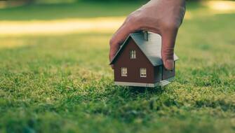 1148812518 Concept of selling a house. A hand is holding a model house above green meadow.