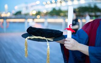 happy proud PhD graduated male student in Academic dress gown holds Graduation cap or hat and diploma or certificate. Blurred night colorful city ligh