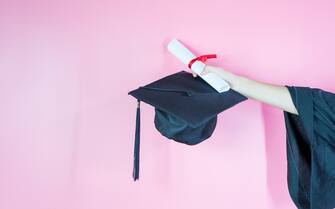 A hand holding graduation cap and diploma on pink background.