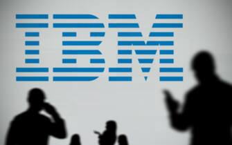 The IBM logo is seen on an LED screen in the background while a silhouetted person uses a smartphone in the foreground (Editorial use only)