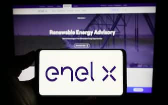 Person holding cellphone with logo of Italian energy company Enel X on screen in front of business webpage. Focus on phone display.