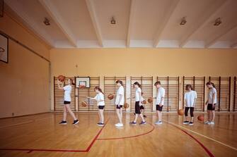 Group of teenage girls wearing white t-shirts and black shorts practicing basketball throws, standing in a row in gym. Wide angle view.
