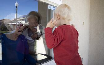 VENTURA, CA - APRIL 3: Mary-Lou McCullagh, 83, and her husband Bob, 84, greet Axel Stirton, 2, the little boy who lives across the street April 3, 2020 in Ventura, California. Mary-Lou and Bob are in isolation from the Covid-19 pandemic, trying to ensure that they do not come in contact with the virus. Mary-Lou has a medical condition that makes her especially vulnerable. They are using Zoom technology to stay in touch with family and friends. Axel is a regular visitor in normal times, but he can no longer come into the house because of the risks of the Covid-19 virus. Mary-Lou and Bob comfort him in his confusion through the window. Mary-Lou is wearing Happy Birthday glasses in honor of her sons birthday which was yesterday. As infection rates soar in the USA, more and more elderly people are in isolation and cut off from any physical contact with their families and friends. (Photo by Brent Stirton/Getty Images)