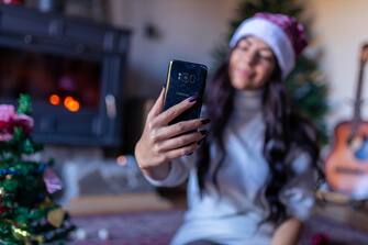 Serene and carefree young woman, taking selfie with her mobile phone, while wearing Santa hat, and celebrating Christmas and New Year holidays in her , well decorated home