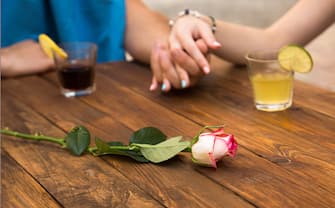 Vintage natural wood cafe table with beverage glasses rose flower on foreground defocused lovers holding hands of each other young male female vacation style environment