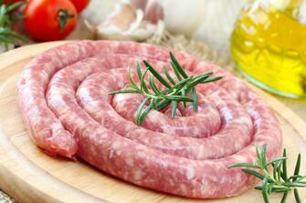 176439643 Raw Italian long pork sausage - LUGANEGA - on a wooden cutting board with olive oil and vegetables at the background ready to be cooked