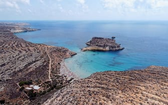 Aerial drone. Spiaggia and Isola dei Conigli, Lampedusa. 
Tranquil, cove-style beach with white sand and turquoise surf bordered by rocky cliffs.