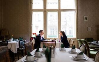 A mature couple is sitting in a romantic old hotel restaurant in front of a big, bright window and having breakfast. They are looking at each other happily and seem to be in love. The rest of the picture shows an old-school, cozy interior.