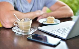 Young guy freelancer working on laptop in cafe.  on the table there is cup of cappuccino coffee, piece of cake on saucer, smartphone