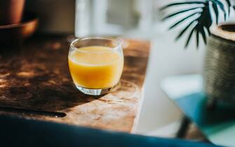 944228702 A single glass of fresh orange juice, sitting on top of a dark wood table in morning sunlight.