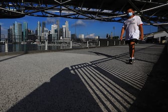 1215155896 SINGAPORE - MAY 26: A man wearing protective mask walks under the Helix Bridge with the central business district pictured in the background on May 26, 2020 in Singapore. Singapore is set to ease the partial lockdown measures against the coronavirus (COVID-19) pandemic after 1 June in three phases to resume activities safely after it sees a decline in the new infection cases in the community. Singapore's gross domestic product (GDP) is expected to shrink as much as 7 percent this year as the country battles the slump in global trade and travel amid the coronavirus pandemic, according to the government report in the local media today. (Photo by Suhaimi Abdullah/Getty Images)