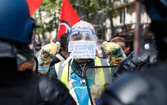 A protester wearing a yellow vest, oven mitts and a protective visor with a sign reading "500,000 euros of tableware, that's a lot of masks Brigitte"  faces gendarmes during a demonstration in Paris, on July 14, 2020, as part of a nationwide day of protests by health workers to demand better work conditions. - Health care workers are protesting in France on the country's National day to demand more for their sector a day after the government and unions signed an agreement giving over eight billion euros in pay rises, with the prime minister admitting the move was overdue in view of the coronavirus pandemic. (Photo by Zakaria ABDELKAFI / AFP) (Photo by ZAKARIA ABDELKAFI/AFP via Getty Images)