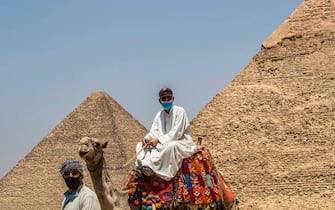 A mask-clad camel guide rides a camel near the (L to R) Great Pyramid of Khufu (Cheops) and Pyramid of Khafre (Chephren) at the Giza Pyramids necropolis on the southwestern outskirts of the Egyptian capital Cairo on July 1, 2020 as the archaeological site reopens while the country eases restrictions put in place due to the COVID-19 coronavirus pandemic. - A spree of openings in Egypt comes after the country officially ended a three-month nighttime curfew a few days earlier. Cafes and shops have re-opened but public beaches and parks remain closed as part of measures to curb the spread of the novel coronavirus. Egypt has recorded more than 65,000 COVID-19 cases including over 2,700 deaths. (Photo by Khaled DESOUKI / AFP) (Photo by KHALED DESOUKI/AFP via Getty Images)