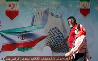 epa09023589 An Iranian couple wearing face masks walks next to the Iranian national flag poster in Tehran, Iran, 19 February 2021. One year has passed since Iran officially announced first positive infection of coronavirus Covid-19. According to Iranian health ministry, 59,341 people have died due to COVID-19 disease since the coronavirus crisis started in the country.  EPA/ABEDIN TAHERKENAREH