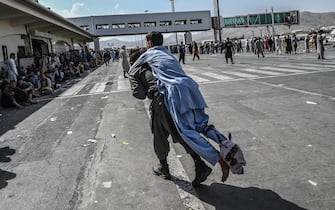1234712990 TOPSHOT - A volunteer carries an injured man as other people can be seen waiting at the Kabul airport in Kabul on August 16, 2021, after a stunningly swift end to Afghanistan's 20-year war, as thousands of people mobbed the city's airport trying to flee the group's feared hardline brand of Islamist rule. (Photo by Wakil Kohsar / AFP) (Photo by WAKIL KOHSAR/AFP via Getty Images)
