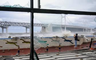 A man standing amongst debris on a road is seen beyond the broken windows (foreground) of a convenience store, while taking photos of waves in front of the Gwangandaegyo or Diamond Bridge (back), after Typhoon Hinnamnor passed through Busan on September 6, 2022. (Photo by Anthony WALLACE / AFP) (Photo by ANTHONY WALLACE/AFP via Getty Images)