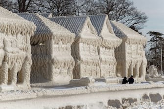 TOPSHOT - People walk by homes covered in ice at the waterfront community of Crystal Beach in Fort Erie, Ontario, Canada, on December 28, 2022, following a massive snow storm that knocked out power in the area to thousands of residents.  (Photo by Cole Burston/AFP) (Photo by COLE BURSTON/AFP via Getty Images)