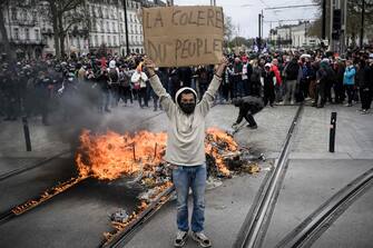 TOPSHOT - A protestor standing beside a fire in the street holds a placards reading "The anger of the people" as protesters face police on the sidelines of a demonstration as part of a national day of strikes and protests, a week after the French government pushed a pensions reform through parliament without a vote, using the article 49.3 of the constitution, in Nantes, western France, on March 23, 2023. - French unions on March 23 staged a new day of disruption against the president's pension reform after he defiantly vowed to implement the change, which includes raising the age of retirement from 62 to 64, saying he was prepared to accept unpopularity in the face of sometimes violent protests. (Photo by LOIC VENANCE / AFP)