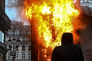 TOPSHOT - A protester wearing a hood stands next to a burning new kiosk beside the Place de l'Opera on the sidelines of a demonstration as part of a national day of strikes and protests, a week after the French government pushed a pensions reform through parliament without a vote, using the article 49.3 of the constitution, in Paris, on March 23, 2023. - French unions on March 23 staged a new day of disruption against the president's pension reform after he defiantly vowed to implement the change, which includes raising the age of retirement from 62 to 64, saying he was prepared to accept unpopularity in the face of sometimes violent protests. (Photo by Alain JOCARD / AFP)