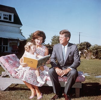 Jacqueline Kennedy and her husband Senator John F. Kennedy sit on a lounge chair at their summer home. Jacqueline reads to her daughter Caroline from a book.