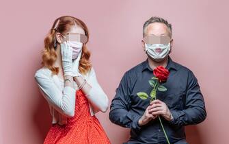 Studio photograph of couple wearing PPE masks, for Covid-19 Valentine's Day. One pink background. Man is holding a red rose, with short greying hair and glasses. Woman has long red hair, wearing a red dress with white sweater and white gloves. Man has lipstick on his PPE mask.
