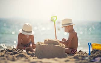 Brothers wearing funny fedora hats having fun building a sandcastle on the beautiful majorcan beach beach. The boys are aged 4. Sunny summer day.