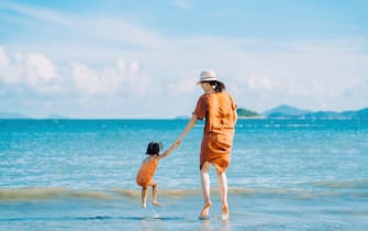 Rear view of joyful young Asian mother and little daughter holding hands while jumping on the beach against blue sky on a sunny Summer day