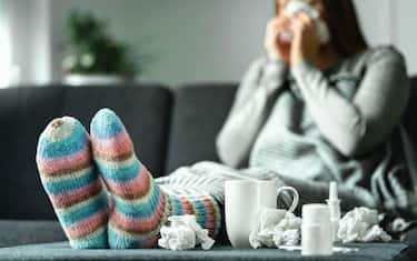Sick woman with flu, cold, fever and cough sitting on couch at home. Ill person blowing nose and sneezing with tissue and handkerchief. Woolen socks.