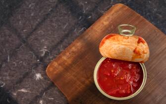 Opened can of arrabbiata sauce, or sugo all'arrabbiata with tomato cubes. Ingredients are tomato juice, tomato cubes, chill, basil, oregano, salt, olive oil and citric acid. Can on wooden board. Black decorative surface. Light effect. High point of view.