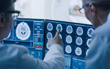 In Control Room Doctor and Radiologist Discuss Diagnosis while Watching Procedure and Monitors Showing Brain Scans Results, In the Background Patient Undergoes MRI or CT Scan Procedure.