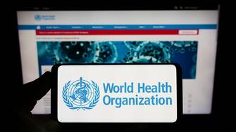 Person holding smartphone with logo of UN agency World Health Organization (WHO) on screen in front of website. Focus on phone display.