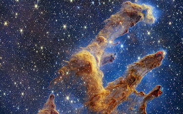 The Pillars of Creation are set off in a kaleidoscope of color in NASA’s James Webb Space Telescope’s near-infrared-light view. The pillars look like arches and spires rising out of a desert landscape, but are filled with semi-transparent gas and dust, and ever changing. This is a region where young stars are forming – or have barely burst from their dusty cocoons as they continue to form.
Newly formed stars are the scene-stealers in this Near-Infrared Camera (NIRCam) image. These are the bright red orbs that sometimes appear with eight diffraction spikes. When knots with sufficient mass form within the pillars, they begin to collapse under their own gravity, slowly heat up, and eventually begin shining brightly.
Along the edges of the pillars are wavy lines that look like lava. These are ejections from stars that are still forming. Young stars periodically shoot out supersonic jets that can interact within clouds of material, like these thick pillars of gas and dust. This sometimes also results in bow shocks, which can form wavy patterns like a boat does as it moves through water. These young stars are estimated to be only a few hundred thousand years old, and will continue to form for millions of years.
Although it may appear that near-infrared light has allowed Webb to “pierce through” the background to reveal great cosmic distances beyond the pillars, the interstellar medium stands in the way, like a drawn curtain.
This is also the reason why there are no distant galaxies in this view. This translucent layer of gas blocks our view of the deeper universe. Plus, dust is lit up by the collective light from the packed “party” of stars that have burst free from the pillars. It’s like standing in a well-lit room looking out a window – the interior light reflects on the pane, obscuring the scene outside and, in turn, illuminating the activity at the party inside.
Webb’s new view of the Pillars of Creation will help researchers revamp models of star formation. By identifying far more precise star populations, along with the quantities of gas and dust in the region, they will begin to build a clearer understanding of how stars form and burst out of these clouds over millions of years.
The Pillars of Creation is a small region within the vast Eagle Nebula, which lies 6,500 light-years away.
Webb’s NIRCam was built by a team at the University of Arizona and Lockheed Martin’s Advanced Technology Center.