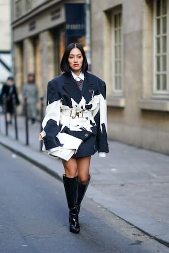 1202323612 PARIS, FRANCE - JANUARY 22: Tiffany Hsu wears a dark blue and white oversized jacket/dress, a white belt, a white clutch, black thigh high boots, outside Maison Margiela, during Paris Fashion Week - Haute Couture Spring/Summer 2020, on January 22, 2020 in Paris, France. (Photo by Edward Berthelot/Getty Images )