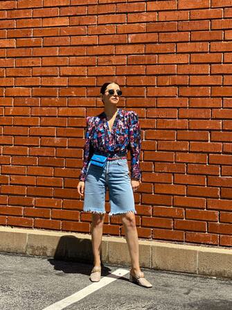 PARIS, FRANCE - APRIL 09: Julia Comil wears a retro floral printed draped top / shirt with padded shoulders by Iro Paris, 90â  s high rise loose fit Bermuda denim Shorts by AGolde, a blue chain belt bag in leather and gold metal by Chanel, cream leather loafers with gold chain Neil J Rodgers, during an online remote street style fashion photo session via Apple iphone / Facetime and the CLOS app as the model is based in Los Angeles - California and the photographer in Paris - France, on April 09, 2021 in Paris, France. (Photo by Edward Berthelot/Getty Images)