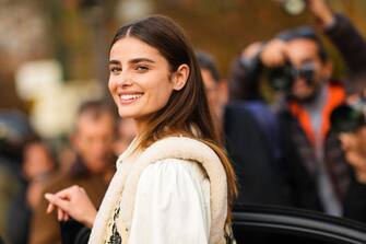 PARIS, FRANCE - OCTOBER 03: Taylor Hill wears gold earrings, a white denim shirt, a beige with black embroidered flower print pattern with sheep interior sleeveless gilet, black with print pattern large pants, a black shiny leather shoulder bag , outside Rokh, Paris Fashion Week - Womenswear Spring/Summer 2023, on October 03, 2022 in Paris, France. (Photo by Edward Berthelot/Getty Images)