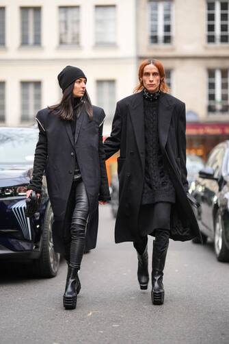 PARIS, FRANCE - JANUARY 19: A guest (L) wears a black wool beanie, a black turtleneck pullover, a black shoulder-pads long coat, black shiny leather legging pants, black shiny leather platform heels ankle shoes, a black braided leather shiny leather handbag, silver rings, a guest (R) wears silver earrings, a black shiny leather nailed / studded large necklace, a black braided wool pullover, a black long coat, a black ribbed wool knees skirt, black legging pants, black shiny leather platform soles and heels knees boots , outside Rains, during Paris Fashion Week - Menswear Fall-Winter 2023-2024, on January 19, 2023 in Paris, France. (Photo by Edward Berthelot/Getty Images)