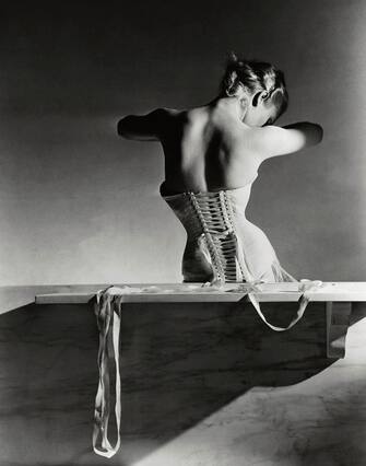 Model, seen from behind, sitting on a wooden bench, looking down through her arms, and wearing a back-lacing corset by Detolle for Mainbocher. (Photo by Horst P. Horst/Conde Nast via Getty Images)