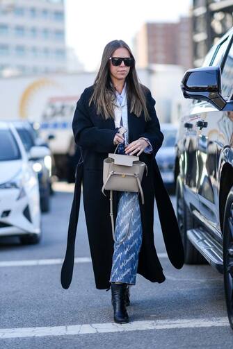 NEW YORK, NEW YORK - FEBRUARY 15: A guest wears black sunglasses, a baby blue and white striped print pattern shirt, a silver shiny leather crocodile print pattern jacket, a black long coat, a gray shiny leather Belt handbag from Celine, blue faded with white inscriptions print pattern large denim jeans pants from Balenciaga, black shiny leather block heels ankle boots, a silver large diamond ring , outside Gabriela Hearst , during New York Fashion Week, on February 15, 2022 in New York City. (Photo by Edward Berthelot/Getty Images)