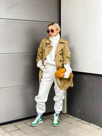 PARIS, FRANCE - APRIL 14: Justyna Czerniak @justynaczerniak_ wears aviator sunglasses from Bottega Veneta, a white wool turtleneck pullover from The Bazilika, a beige trench coat from Dorothee Schumacher, white sport jogger pants from The Bazilika, a brown leather pouch golden chain Bottega Veneta bag, green ankle Nike Jordan Mr Kicks sneakers shoes, during an online remote fashion photo session via Apple iphone / Facetime and the CLOS app as the model is based in Poznan - Poland and the photographer in Paris - France, on April 14, 2021 in Paris, France. (Photo by Edward Berthelot/Getty Images)