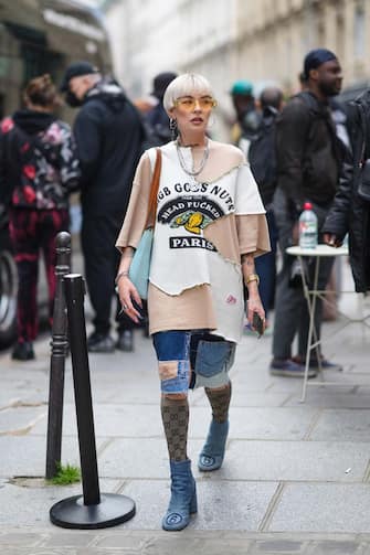 PARIS, FRANCE - JUNE 23: A guest wears yellow sunglasses, silver and gold earrings, a silver chain necklace with a GG pendant from Gucci, a silver chain necklace, a beige with white ripped oversized t-shirt sweater with black print slogan and logo pattern from 8-IGB, a gold watch, gold rings, a silver chain bracelet, silver rings, a pale blue shiny leather embossed Louis Vuitton monogram shoulder bag from LV, blue faded / navy blue patched ripped shorts, brown and black GG monogram knees socks from Gucci, blue faded denim block heels ankle boots with a navy blue 6 seams on the toe-cap, outside BLUEMARBLE, during Paris Fashion Week - Menswear Spring/Summer 2022, on June 23, 2021 in Paris, France. (Photo by Edward Berthelot/Getty Images)