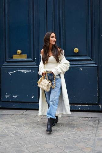 1316671415 PARIS, FRANCE - APRIL 30: Tamara Kalinic wears gold earrings, a gold chain pendant necklace, a white ribs bustier Khaite top with long sleeves, white long oversized Magda Butrym coat, blue faded large oversized Margiela denim jeans pants, a beige suede Bottega Veneta Cassette handbag, black shiny leather pointed heels western Khaite ankle boots, on April 30, 2021 in Paris, France. (Photo by Edward Berthelot/Getty Images)