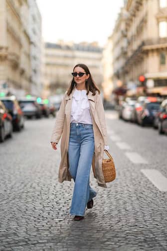 PARIS, FRANCE - MARCH 02: Marielle Haon wears sunglasses, a white ruffle shirt, a beige trench long coat, blue denim flare jeans pants, a brown wicker basket bag, burgundy leather shoes, on March 02, 2021 in Paris, France. (Photo by Edward Berthelot/Getty Images)