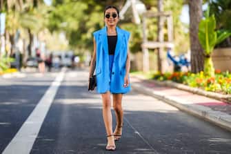 CANNES, FRANCE - MAY 19: Nesrine Bouazzaoui wears black circle sunglasses from Ray Ban, silver earrings, a black shoulder-off / sleeveless t-shirt, a flashy blue sleeveless / long blazer jacket, matching blue flashy suit shorts, a brown LV monogram print pattern zipper clutch from Louis Vuitton, gold bracelets, gold rings, black shiny leather strappy heels sandals, on May 19, 2022 in Cannes, France.  (Photo by Edward Berthelot / Getty Images)