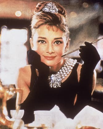 Belgian-born actress Audrey Hepburn (1929 - 1993), in a black, shoulderless dress, matching gloves, and a tiara, smiles with a cigarette holder in her hand, in her role as Holly Golightly the film, 'Breakfast at Tiffany's,' directed by Blake Edwards, New York, New York, 1961. (Photo by Paramount Pictures/Courtesy of Getty Images)