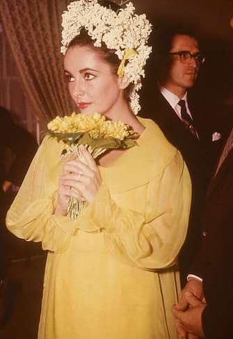 British-born actor Elizabeth Taylor, a yellow dress and floral headdress, holds a bouquet of flowers at her wedding to actor Richard Burton, March 15, 1964. (Photo by Hulton Archive/Getty Images)