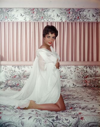 British-born actor Elizabeth Taylor kneels on a bed and wears a sheer white nightgown and robe, circa 1950s. (Photo by Hulton Archive/Getty Images)