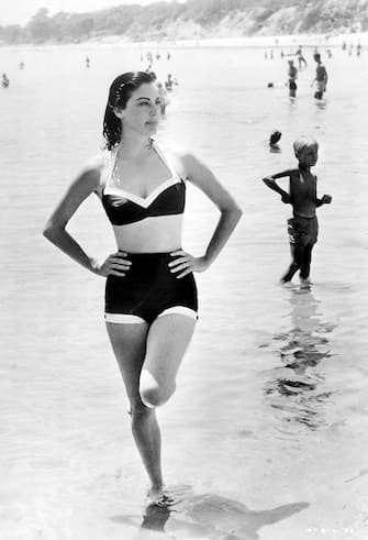 Actress Ava Gardner on the beach.  She wears a two-piece bathing suit and stands with one foot up in ankle-deep water.  Undated photograph.