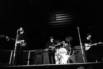 August 1964: The Beatles in concert in the United States.  The Stars and Stripes flag is illuminated in the background.  (Photo by Express / Express / Getty Images)