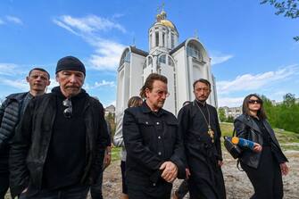 Andrii Holovine, priest of the Church of St. Andrew Pervozvannoho All Saints (2nd,R) guides Bono (C) (Paul David Hewson), activist and front man of the Irish rock band U2 and guitarist David Howell Evans aka 'The Edge' (R) visiting the site of a mass grave by the church in the Ukrainian town of Bucha, near Kyiv on May 8, 2022. (Photo by Genya SAVILOV / AFP) (Photo by GENYA SAVILOV/AFP via Getty Images)