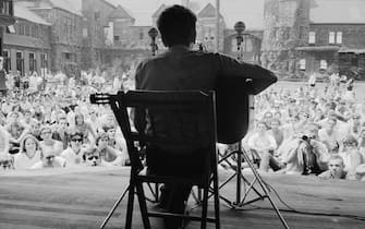 View, from behind, of American musician Bob Dylan as he plays acoustic guitar during a performance at the Newport Folk Festival, Newport, Rhode Island, July 1963. Audience members sit on the grass of the tennis court at the ivy-covered buildings of the Newport Casino (later the National, and then International, Tennis Hall of Fame). (Photo by Rowland Scherman/Getty Images)